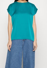 Maglietta corta turchese con scollo rotondo, texture liscia e vestibilità rilassata; abbinata a jeans in denim blu a gamba larga.