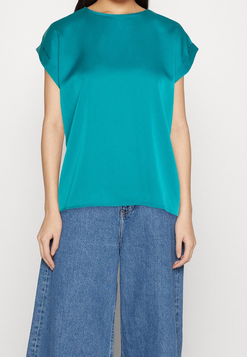 Maglietta corta turchese con scollo rotondo, texture liscia e vestibilità rilassata; abbinata a jeans in denim blu a gamba larga.