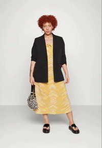 Blazer preto sobre um vestido amarelo com padrão e detalhe de laço. Usado com sandálias pretas e segurando uma bolsa com riscas. Cabelo curto e cacheado vermelho.