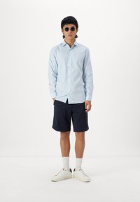 Lichtblauw overhemd met lange mouwen en knopen, marineblauwe shorts, witte sneakers en ronde zonnebril; casual stijl, soepele stof en effen kleuren.