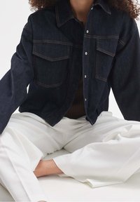 Person som bär en mörk denimjacka över en brun topp och lösa vita byxor, sittande med korslagda ben mot en enfärgad bakgrund.