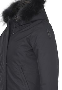 Giacca nera isolata con bordo in finta pelliccia sulla cappuccio, chiusura con zip, tasche laterali e un sottile logo ricamato sulla manica.