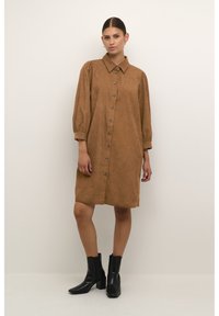 Robe chemise en velours côtelé marron avec un devant à boutons, un col et des manches bouffantes. Associée à des bottines noires, créant un contraste de couleurs et de textures.