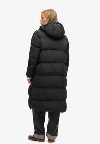Sort pufferjakke med hætte, med en quiltet design. Lang, oversized pasform, glat tekstur og synlig lynlås på siden.