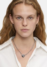Silverhalsband med hänglåsberlock. Kedjan har stora länkar och hänglåset har en svart detalj. Bakgrunden är neutral.