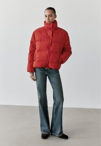 Rote Steppjacke mit hohem Kragen und Knopfverschluss. Getragen mit ausgestellten blauen Jeans und schwarzen Schuhen. Glatte Textur, warmer Eindruck.