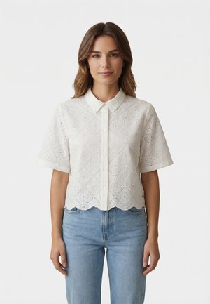 Donna con capelli castani di media lunghezza che indossa una camicia bianca a maniche corte con occhielli e bottoni, e jeans azzurri, in piedi davanti a uno sfondo bianco.