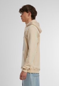 Beige hættebluse med en frontlomme, snor i hætten og blød tekstur. Modellen har lyseblå jeans på; profilbillede mod en neutral baggrund.