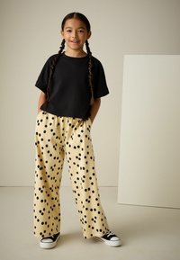 Camiseta negra corta, pantalones amarillos con lunares negros, de pie con las manos en los bolsillos, zapatillas negras, cabello largo trenzado.