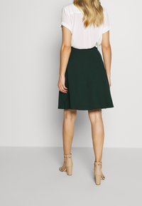 Donna che indossa una blusa bianca a maniche corte, una gonna verde scuro fino al ginocchio e sandali con tacco a blocco beige, in piedi contro uno sfondo neutro.