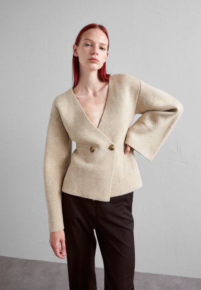 Beige Wollmischung-Wickeljacke mit tiefem V-Ausschnitt, doppelter Knopfleiste und weiten Ärmeln. Kombiniert mit schwarzen Hosen. Glatte Textur.