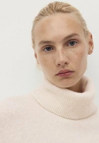 Svetlo roza pleteni pulover s turtleneck ovratnikom, z rebrastim ovratnikom in mehko teksturo, ki prikazuje bližnji posnetek obraza nosilca.
