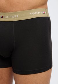 Κοντινό πλάνο ενός ατόμου που φοράει μαύρα εσώρουχα τύπου boxer με μπεζ ζώνη μέσης που φέρει το λογότυπο και τη σημαία της Tommy Hilfiger.