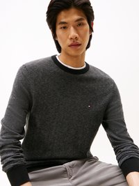 Maglione grigio a motivo a spina di pesce con collo tondo a costine nero e polsini. Piccolo logo ricamato sul petto. Modello che indossa pantaloni grigi casual.