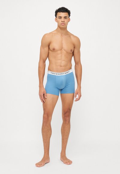 Fiatal férfi áll mezítláb fehér háttér előtt, világoskék boxeralsót visel fehér Polo Ralph Lauren derékrésszel.