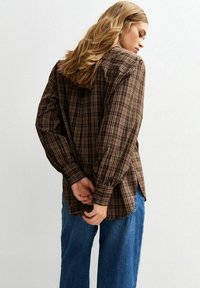 Brown plaid shirt with long sleeves, button cuffs, and side slits, paired with blue wide-leg jeans, model facing away from the camera.
