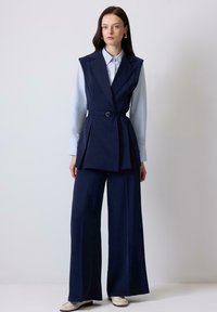 Navy blue tailored vest with a waist belt, layered design, and wide-legged trousers in matching fabric. Light blue collared shirt underneath.