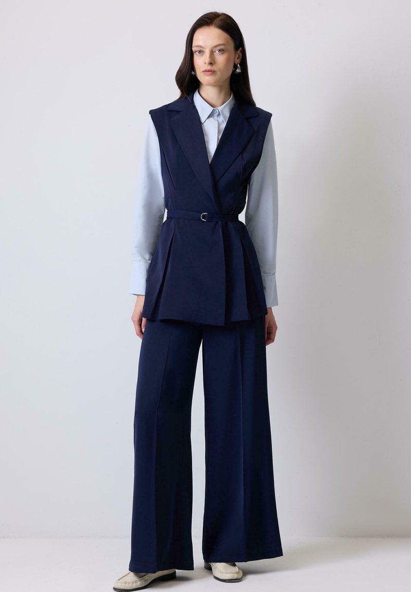 Navy blue tailored vest with a waist belt, layered design, and wide-legged trousers in matching fabric. Light blue collared shirt underneath.