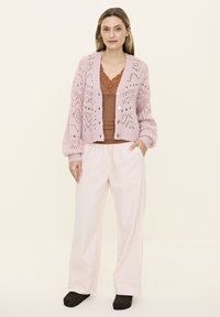Light pink knitted cardigan with a diamond pattern, paired with a brown tank top and loose-fitting cream pants, worn with black shoes.