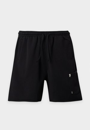 Mann steht mit Händen in den Taschen, trägt ein schlichtes weißes T-Shirt, schwarze Shorts, weiße Socken und schwarze Sneaker auf weißem Hintergrund.