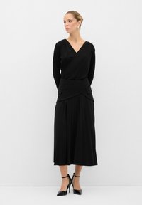 Robe noire avec un décolleté en V, des manches longues et une jupe plissée. Présente des poches accentuées, une texture lisse et est assortie à des escarpins noirs.
