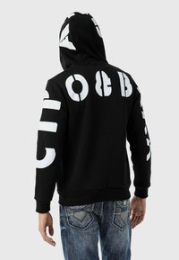 Schwarzer Kapuzenpulli mit großen weißen Buchstaben auf dem Rücken und den Ärmeln. Hergestellt aus einem Baumwollmix mit strukturierter Oberfläche.