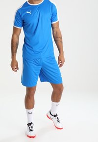 Blå idrottströja med vita detaljer, tillsammans med blå shorts. Har Puma-logotyp. Bärs med vita strumpor och sportiga skor.