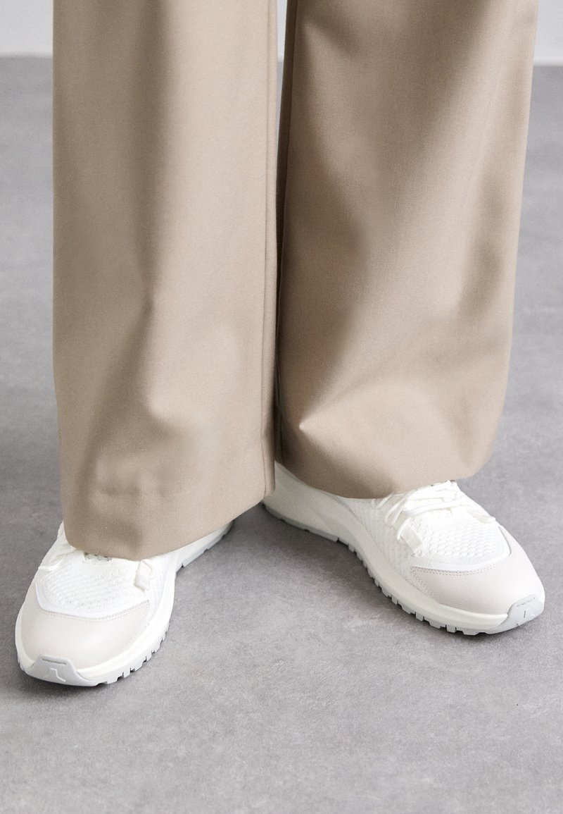 Valkoiset tennarit, joissa on verkkokangasta ja sileitä osia, varustettuna teksturoidulla pohjalla, yhdistettynä beigeihin leveisiin housuihin. Näkyvät nauhat ja yksityiskohdat.