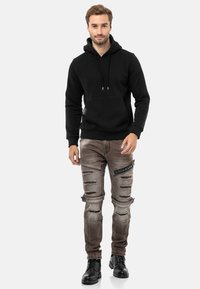 Schwarzer Hoodie mit Kängurutasche und Kordelzügen, kombiniert mit ausgewaschenen grauen Jeans mit Rissen und einem seitlichen Patch. Schwarze Stiefel runden das Outfit ab.