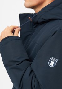 Chaqueta con capucha de color azul marino con un acabado texturizado, que presenta un parche del logo en la manga y un cordón ajustable en el cuello.