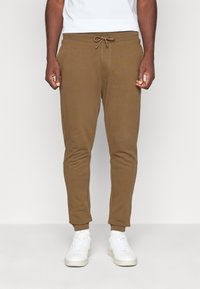 Bruine katoenen joggers met een elastische tailleband en trekkoord, met taps toelopende benen en geribde manchetten, gedragen met witte sneakers.