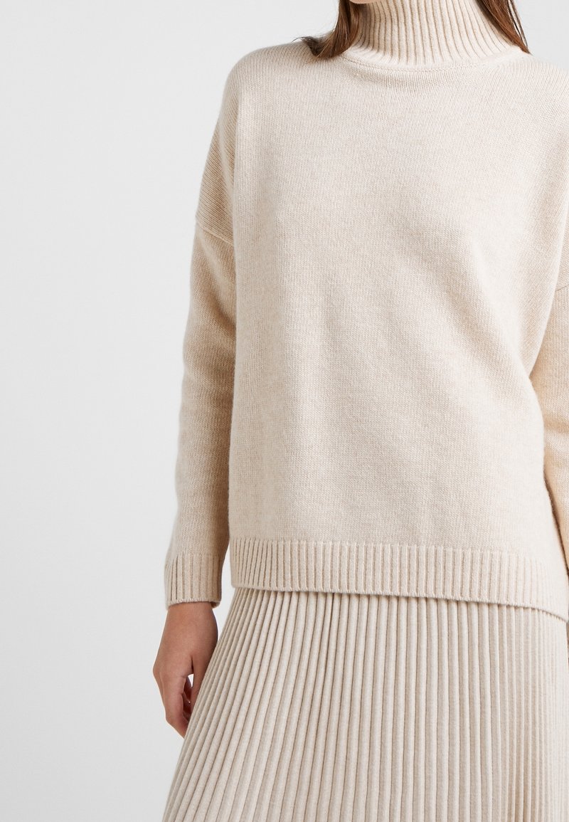 Femme portant un pull à col roulé côtelé beige et une jupe plissée beige assortie sur un fond uni.