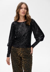 Vrouw in een zwarte pailletten top met lange mouwen en opengeslagen mouwen, met bruine broek met luipaardprint, staand tegen een effen achtergrond.