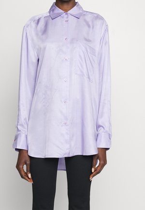 Persona vistiendo una camisa de satén lavanda, holgada y de manga larga, con un bolsillo en el frente y cierre de botones, combinada con pantalones negros.