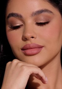 Primer plano de un rostro con piel suave, labios prominentes en un tono rosa suave, sombra de ojos marrón claro y pestañas largas. Mano descansando en la barbilla.