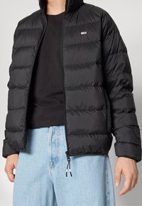Svart puffjacka med dragkedja framtill, horisontellt quiltat mönster och en liten logotyp på bröstet. Bärs över en svart skjorta och ljusa jeans.
