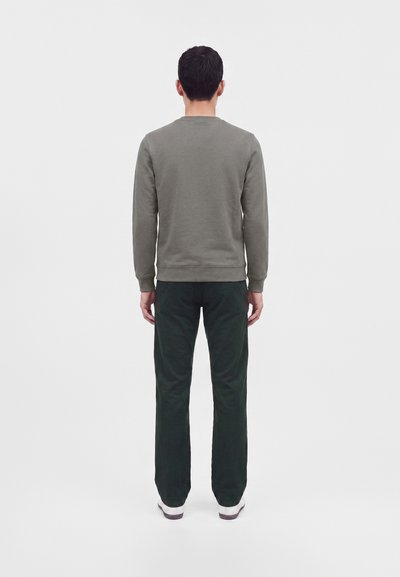 Homme de dos portant un sweat-shirt gris, un pantalon vert foncé et des chaussures blanches sur un fond uni gris clair.