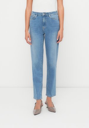 Frau trägt hellblaue Jeans mit geradem Bein, weißes Top und taupefarbene Slingback-Pumps mit spitz zulaufender Spitze, steht vor weißem Hintergrund.