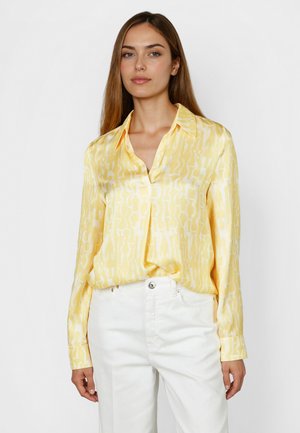 Woman wearing a light yellow patterned silk blouse with white high-waisted pants standing against a plain white background.