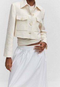 Lyhytnukkeinen kermajakku, jossa on teksturoitu kangas, kaksi rintataskua ja nappikiinnitys, yhdistettynä vaaleanväriseen paitaan ja valkoiseen hameeseen.