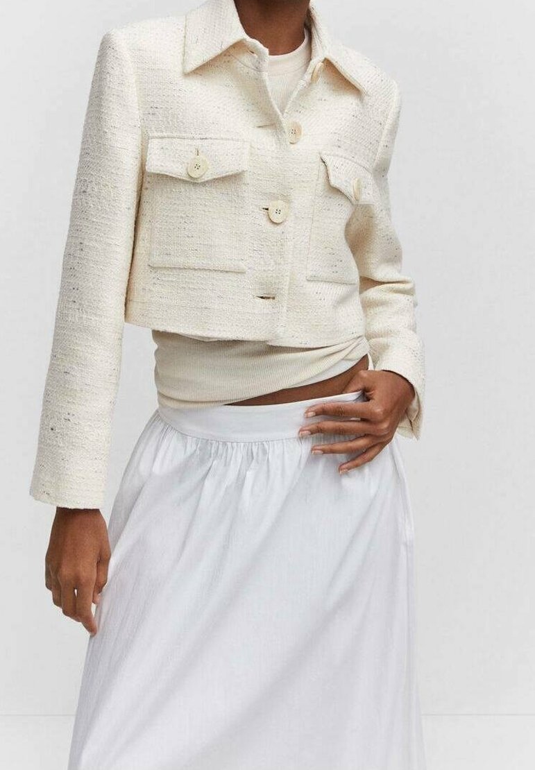 Lyhytnukkeinen kermajakku, jossa on teksturoitu kangas, kaksi rintataskua ja nappikiinnitys, yhdistettynä vaaleanväriseen paitaan ja valkoiseen hameeseen.