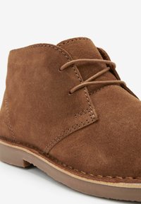 Botín de ante marrón hasta el tobillo con diseño de cordones, con detalles de costura y suela de goma gruesa. Textura suave y forma de punta redondeada.