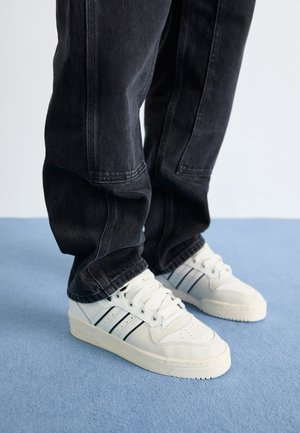 Hvide sneakers med sorte striber båret med løse sorte jeans på et blåt tæppe mod en lys baggrund.