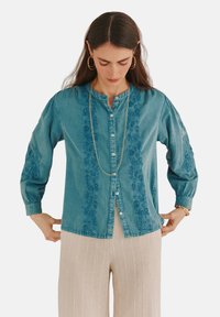 Hellblaues Denimhemd mit Knopfleiste, langen Ärmeln und floralem Stickerei auf der Brust. Kombiniert mit cremegestreiften Hosen.