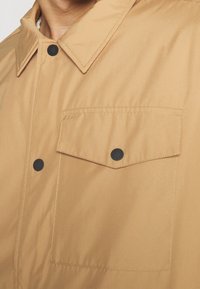 Veste couleur fauve avec col pointu, poche avant à rabat et boutons-pression noirs sur la poche et la patte de boutonnage.