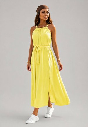 Femme portant une robe longue jaune sans manches avec une ceinture à la taille, des baskets blanches, un foulard marron et des bijoux dorés, debout sur un fond gris.
