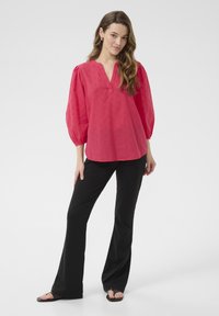 Blouse rouge framboise avec manches ballon, décolleté en V et motif texturé ; associée à un pantalon noir évasé et des tongs noires.