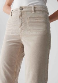 Beige Cord-Hosen mit geradem Schnitt, vorderen Taschen und einer einzelnen Gesäßtasche mit horizontalen Nähten. Sichtbares Stoffdetail mit Textur.