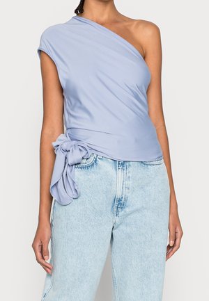 Jasnoniebieski top na jedno ramię wykonany z miękkiego materiału, z kopertowym fasonem i wiązaniem w talii, zestawiony z jasnoniebieskimi jeansami.