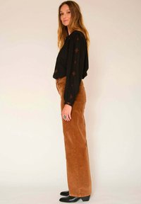 Blouse noire avec broderies rouges, pantalon large en velours côtelé marron, et bottines noires. Vue de profil mettant en avant les détails de la tenue.